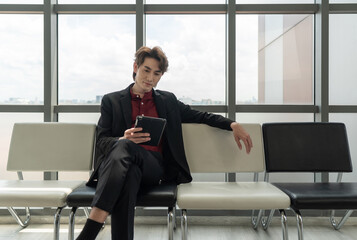 Young asian businessman dressed in a smart suit sit comfortably on a bench, engaging with a tablet...