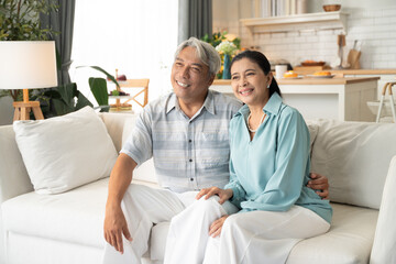 Obraz premium Asian senior man embracing wife on sofa and talking together. Retired couple spending time in the morning at home. Healthy and life insurance concept