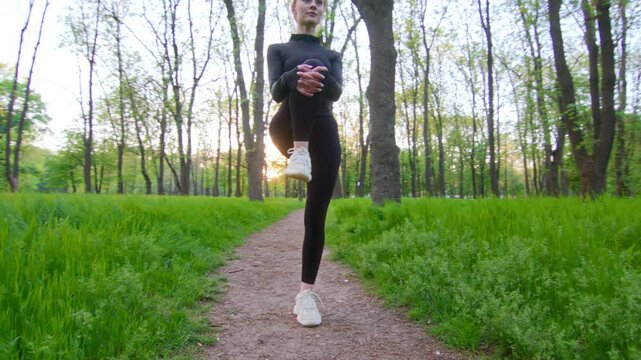 Lifting her knee for ankle rotation, then drawing the foot into a stretch, a blonde woman maintains steady balance. The slow-motion camera arcs around her focused posture amid forest calm.