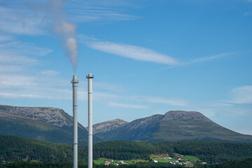 Two industrial chimneys release smoke into the clear blue sky, contrasting with the lush forest, green fields, and rocky mountains of the Norwegian landscape