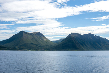 High mountains of Gjevenesstranda nature reserve, along Storfjord in Norway, near Solavagen, under a bright blue sky with scattered clouds seen from the water on a sunny day