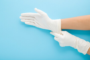 Young adult woman hands wearing white latex protective gloves on light blue background. Pastel...