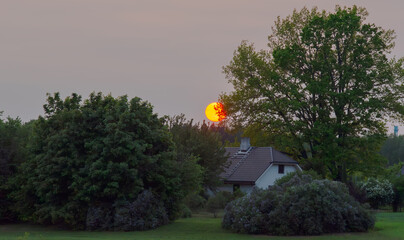 A serene countryside sunset with the orange sun setting behind lush green trees and a cozy white house nestled in the landscape.