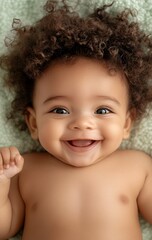A happy baby with curly brown hair smiles broadly while lying on a soft green blanket