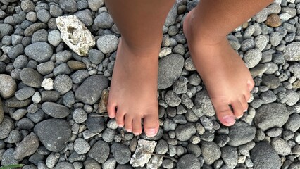 A close-up view shows a child's bare feet standing on a surface covered with numerous grey and brown river stones