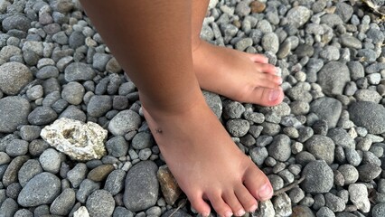 A close-up view shows a child's bare feet standing on a surface covered with numerous grey and brown river stones