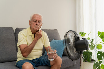 Senior man sitting on sofa wiping sweat from face holding glass of water near electric fan at home
