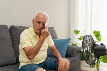 Senior man wiping sweat from face while sitting on sofa near an electric fan at home