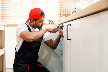 Focused handyman repairing a kitchen cabinet hinge under the sink at home, showcasing plumbing service, repair expertise, home maintenance, and handyman business support for property care.