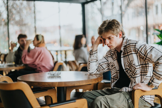 A young man sitting alone in a cafe, appearing thoughtful, surrounded by other engaged patrons.