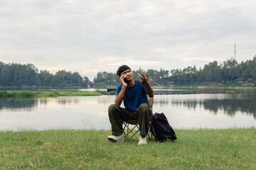 A young man sitting on a chair at lake with relaxing mood,  smiling and engaging in cheerful phone conversation.