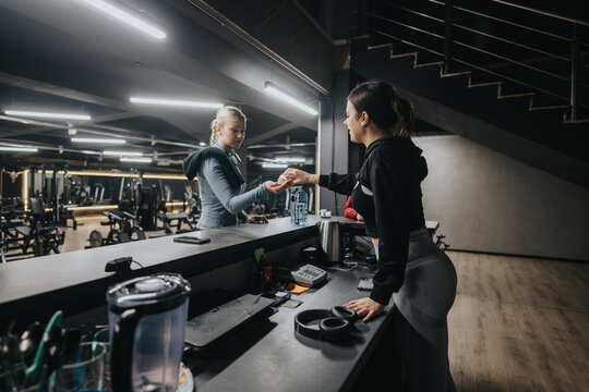 A woman is checking into a gym at the reception desk. They are communicating in a modern fitness center environment, featuring exercise machines and professional equipment.