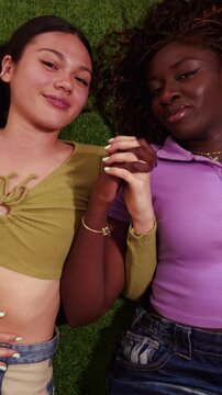 Multi ethnic lesbian partners relaxing on grassy lawn, sharing intimate moment while holding hands and exchanging loving smiles with genuine connection