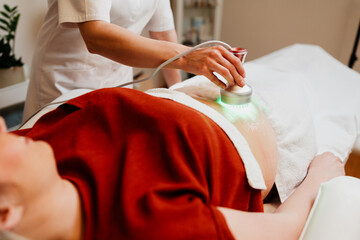 Professional beautician performing a green light therapy treatment on a client's abdomen in a modern beauty spa, promoting skin rejuvenation and collagen production
