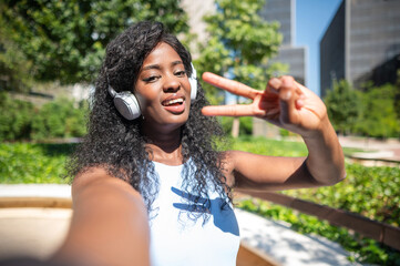Young woman taking selfie while listening music with headphones and making victory sign in city park