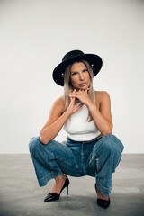 Fashion model in stylish outfit squatting indoors with a serious expression and bold hat