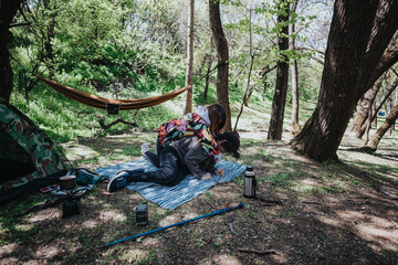 A vibrant image of a young couple spending quality time in a forest. They relax on a blanket next to camping gear, surrounded by nature.