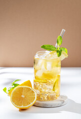Iced tea with lemon and mint in a glass on a white beige background with citrus fruit and sunlight.
