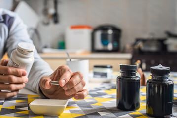 Adult man in home kitchen holding bottle of nutritional supplements, closeup photo. Man sitting at kitchen table holding supplement capsule, bottles and cup in background