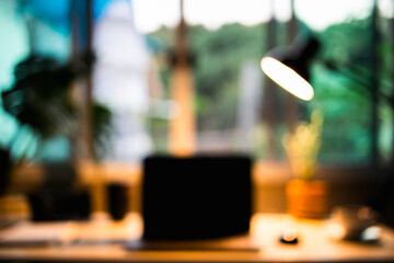 blurred workspace scene with a notebook, digital camera, lamp on  mini desk by the window accented by soft light and an ornamental monstera tree in the background.