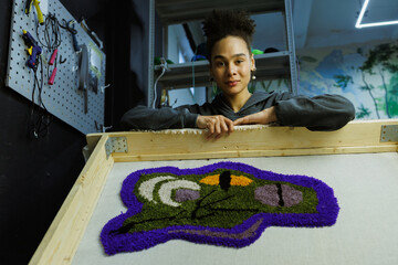 African american craftswoman looking at camera near handmade rug on frame