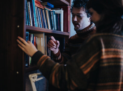 Two friends enjoy a quiet moment indoors as they browse books on a bookshelf, showcasing camaraderie and shared interests while surrounded by a cozy and relaxed atmosphere.