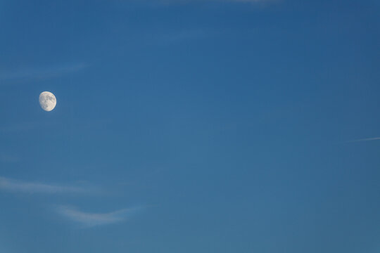 A waxing gibbous moon hangs in a clear blue sky with faint streaks of clouds. The stark contrast highlights the moon's craters against the vast expanse of a blue hour sky, exuding calmness. - Powered by Adobe