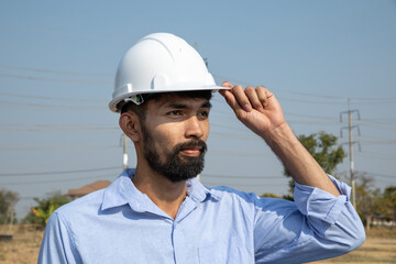 Handsome bearded young engineer wearing a hard hat and vest standing on wind farm.