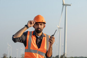 A bearded young engineer wearing a hard hat and holding walkie-talkie stands on a wind farm, looking at camera.