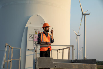 Young bearded engineer or maintenance technician wearing a hard hat using walkie-talkie on a wind farm.