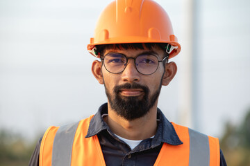 Portrait of handsome bearded young engineer wearing a hard hat and glasses stands on wind farm.