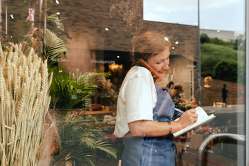 Middle-aged female florist in apron arranging calling on the smart phone. She is arranging...