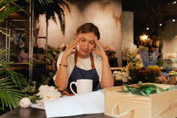 A middle-aged woman calculates finances on a calculator at a flower shop with various plants in the...