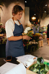 Middle-aged female florist in apron arranging fresh flowers for bouquet in the flower shop. Small...