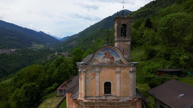 Fixed rone view of a weathered mountain chapel with bell tower and painted fresco, set in lush alpine valley. Shot in Vico, Edolo, Italy (Vico, Edolo, Italia)