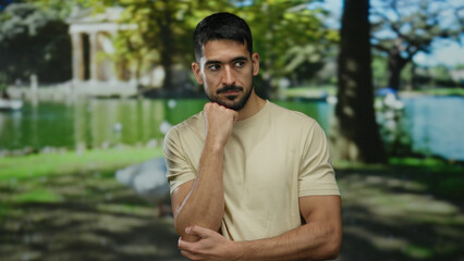 Pensive man outdoors in park setting with thoughtful expression wearing casual beige shirt surrounded by lush trees and peaceful lake under vibrant sunlight