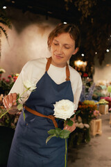 Middle-aged female florist in apron arranging fresh flowers for bouquet in the flower shop. Small...