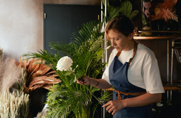 Middle-aged female florist in apron arranging fresh flowers for bouquet in the flower shop. Small...
