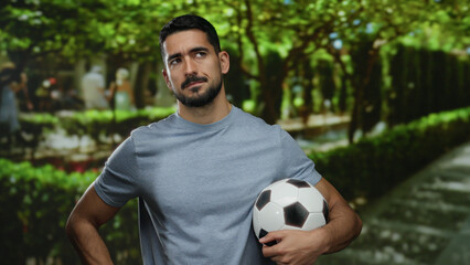 Confident man holding soccer ball outdoors in sunny park setting with lush greenery and blurred people in background, wearing casual gray shirt and sporting beard and short hair
