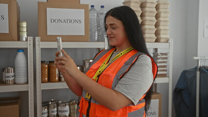 Woman volunteer in a charity center takes a photo with her phone surrounded by donation boxes and food supplies, reflecting a dedicated and supportive community environment.