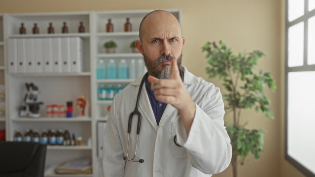 Man with beard and bald head in a clinic wearing a lab coat and stethoscope gestures seriously in a medical room with shelves filled with supplies and a plant nearby.