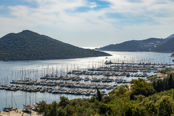 Panoramic view of Kaş Marina with rows of yachts, shimmering water under sun, green hills, and a...
