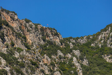 Sleeping Giant summit with Turkish flag and tiny human figure on viewpoint, rocky green mountain landscape. Kaş, Antalya, Mediterranean, Turkey. Sunny spring day.