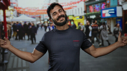 Obraz premium Hispanic man with beard gestures playfully on a busy street with colorful flags, surrounded by blurred crowd in an urban outdoor scene during daytime.