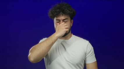 Young man with beard sneezing against blue background, closing eyes and holding nose while wearing...