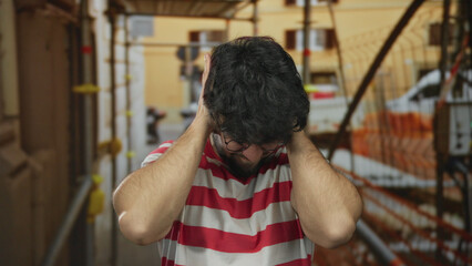 Young man with beard at construction site outdoors appears stressed holding head with hands in...