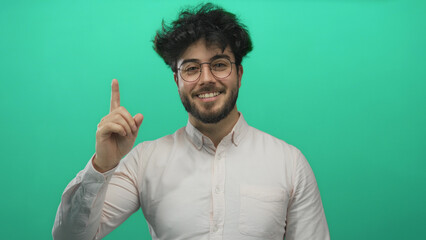 Young man with beard wearing glasses points upward against a vibrant green background, exuding...