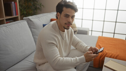 Handsome young hispanic man using phone in cozy living room apartment, wearing casual sweater, looks confident and relaxed indoors, reflecting modern lifestyle and technology.