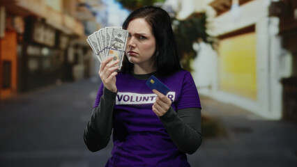 Woman in purple volunteer shirt on street contrasts emotions holding us dollars and credit card,...