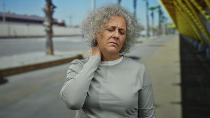 Elderly woman with grey hair touches her neck in discomfort while standing on an urban street,...